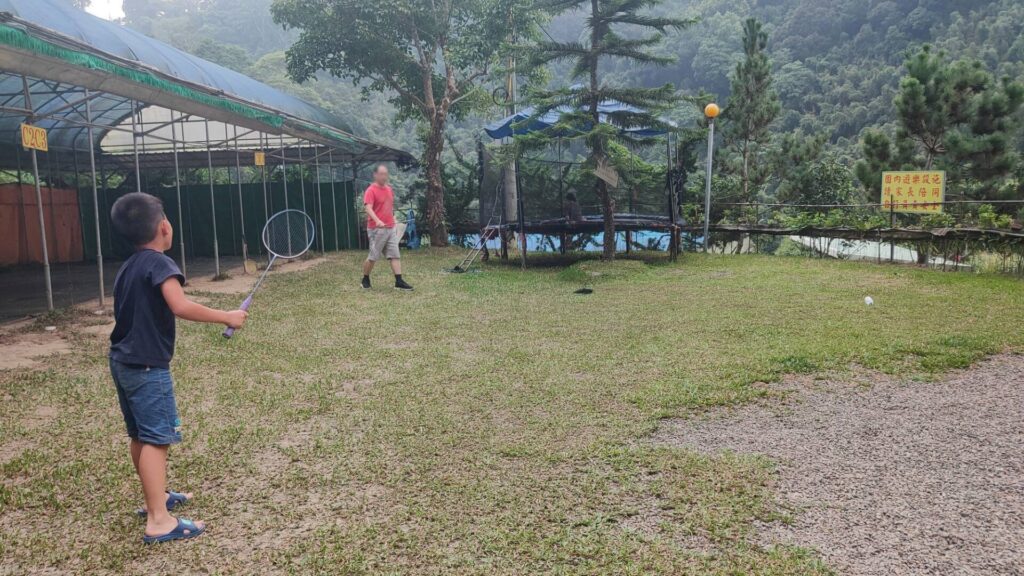 苗栗夢之谷露營區的 C 區雨棚營位前，家人正在寬敞的草地上打羽毛球，背景處可見孩子們最愛的彈跳床等遊樂設施。
